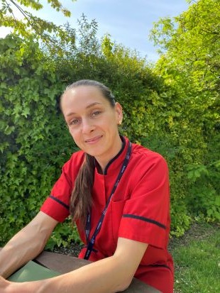 A midwife in red matron tunic smiles, there are trees behind her