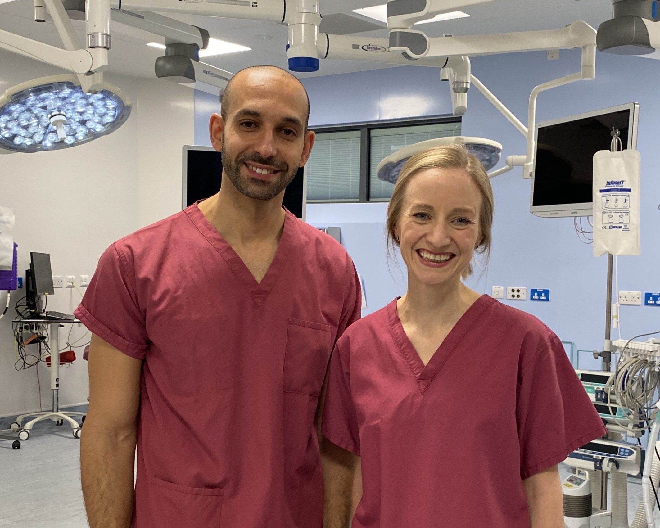 Jess and Islam are stood in a theatre at Southmead Hospital. They are wearing dark pink scrubs and smiling at the camera.