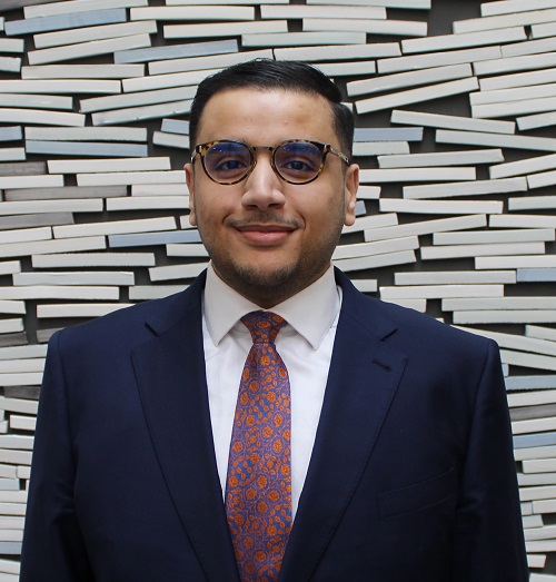 A man with glasses, wearing a suit stands against a tiled wall smiling