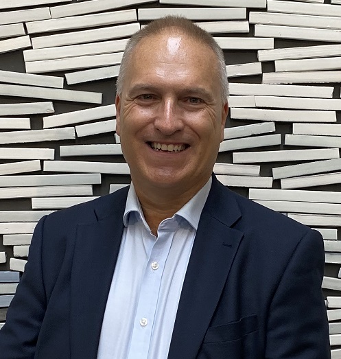 A man in a shirt and jacket stands, smiling, against a tiled wall