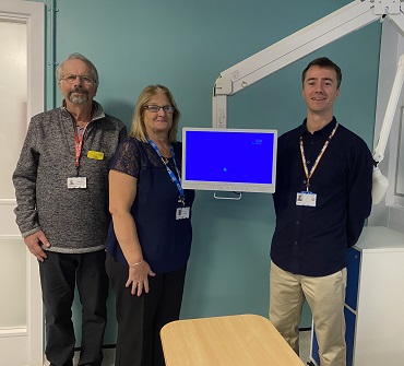 Three people stand together beside a patient bedside TV, smiling at the camera