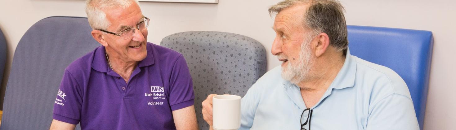 volunteer wearing purple polo shirt chatting with a patient holding a cup of tea