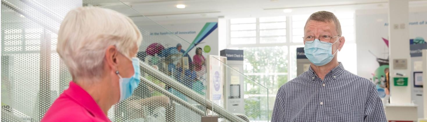 Patient wearing a facemask in the Brunel atrium talking to a volunteer move maker in a pink polo shirt. Patient check-in stands can be seen in the background.