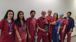 A group of people in raspberry-coloured scrubs and one in blue scrubs, including a man in the middle holding a trophy