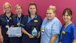 A group of nursing staff in uniform stand in a group, smiling, holding a certificate and trophy 