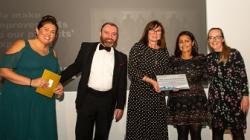 Three women receive a certificate from a man in a tuxedo and a woman in a green dress