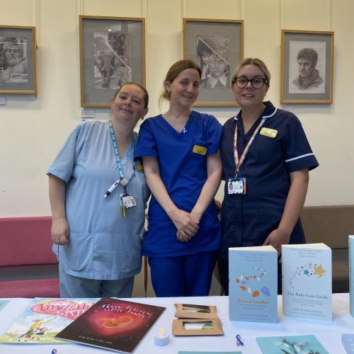 Three members of staff from our Women's and Children's Division in uniform, smiling and standing behind a table with books and information leaflets. 
