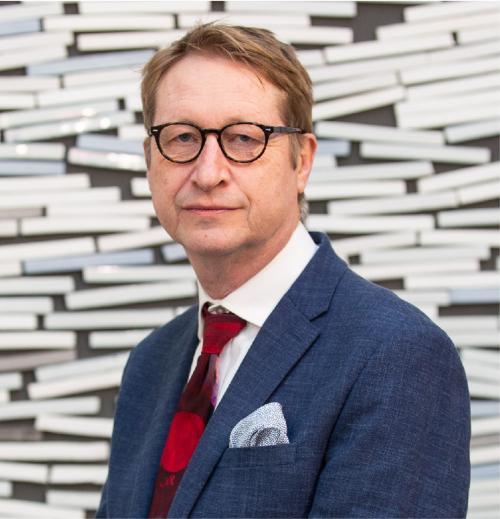 A head and shoulders photo of John looking at the camera. He is wearing a red tie and navy blue suit and glasses.