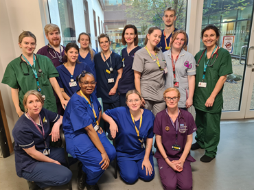 A group of staff in different coloured hospital scrubs stand together in a group smiling