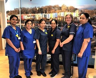 A group of nurses in blue and navy-coloured scrubs stand in front of a mural featuring the colourful houses of Bristol Harbourside, smiling at the camera