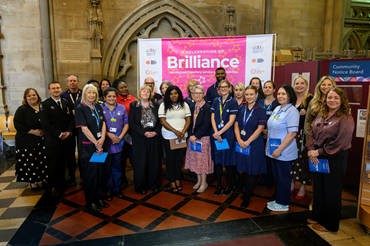 A group of people stand gathered together in front of a bright pink banner that reads Brilliance