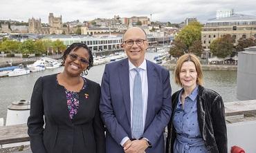 Three people standing on a balcony, smiling at a camera, the Bristol skyline behind them