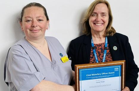 A woman in light grey scrubs is handed a certificate by a person not in NHS uniform. The certificate says: "Chief Midwifery Officer Award".