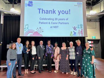 A group of people stand together in front of a sign that says "Thank you! Celebrating 20 years of Patient and Carer Partners at NBT"