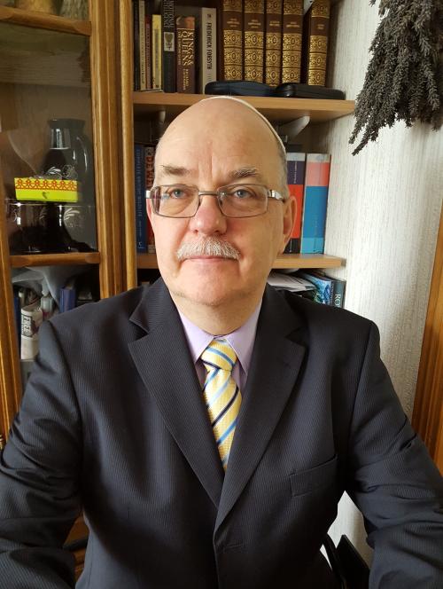 A head and shoulders photo of Jay-Jay. He is wearing a suit and tie, and a pair of glasses. A bookcase can be seen in the background.