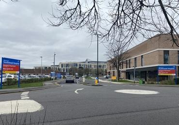 A mini roundabout with two hospital signs, a road and buildings
