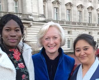 Three people stand outside a grand building (Buckingham Palace)