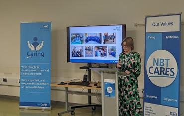 Person stands at a lectern speaking, a presentation showing NHS staff on a screen behind and two blue banners saying "Caring" and "NBT Cares" either side of them.