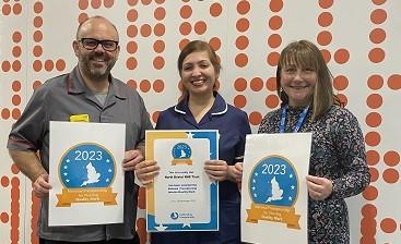 A man in dark grey nurse tunic, a woman in navy nurse tunic and a third woman stand holding certificates, smiling