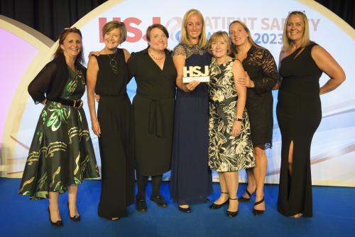 A group of seven people on stage collecting their awards at the HSJ award. They are all in formal dress and the person in the middle of the group is holding the HSJ award.