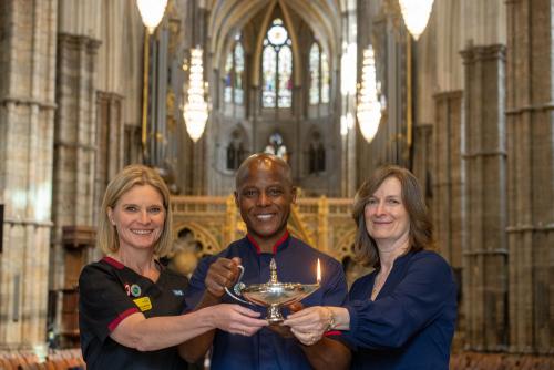 Three people stand together inside a cathedral near ornate wooden choir stalls with gold detailing. Two individuals wear NHS uniforms, and one wears a patterned dress. Other attendees and architectural features, including arches and stained glass, are visible in the background.