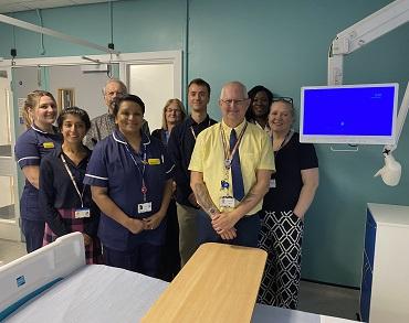 A group of people, some in nursing uniform stand together next to a patient bedside television screen