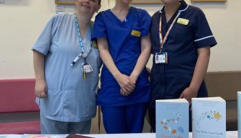 Three members of staff from our Women's and Children's Division in uniform, smiling and standing behind a table with books and information leaflets. 