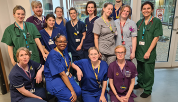 A group of staff in different coloured hospital scrubs stand together in a group smiling