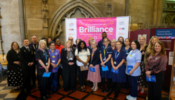 A group of people stand gathered together in front of a bright pink banner that reads Brilliance