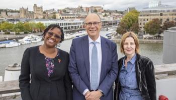 Three people standing on a balcony, smiling at a camera, the Bristol skyline behind them