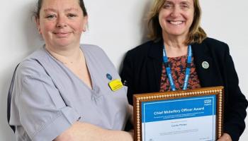A woman in light grey scrubs is handed a certificate by a person not in NHS uniform. The certificate says: "Chief Midwifery Officer Award".