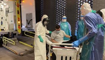 People dressed in PPE covering them from head to toe move a patient from an ambulance trolley to a wheelchair