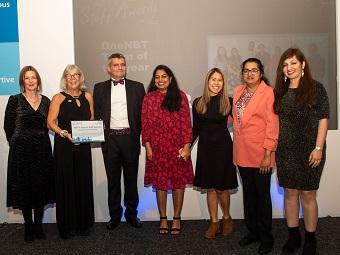 A group of nurses in evening dress stand alongside a man in a tuxedo as they are presented with a framed certificate