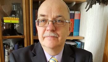 A head and shoulders photo of Jay-Jay. He is wearing a suit and tie, and a pair of glasses. A bookcase can be seen in the background.