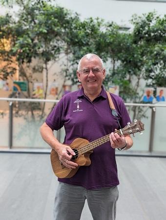 A man in a purple T-shirt holds a ukulele standing, smiling, in front of indoor trees (inside the Brunel atrium, Southmead Hospital)