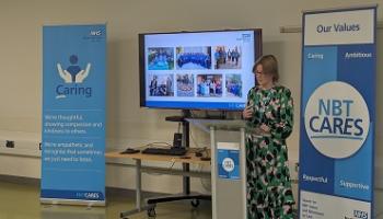 Person stands at a lectern speaking, a presentation showing NHS staff on a screen behind and two blue banners saying "Caring" and "NBT Cares" either side of them.