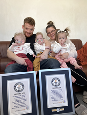 A man and a woman hold three toddlers on their laps. Two large certificates in front of them