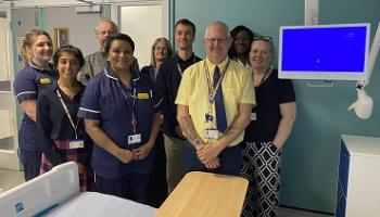 A group of people, some in nursing uniform stand together next to a patient bedside television screen